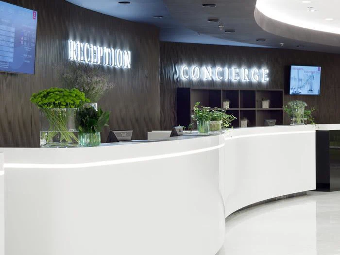En moderne hotelreception med en buet hvid skranke, grønne planter og oplyste skilte med teksten Reception og Concierge på en mørk væg.