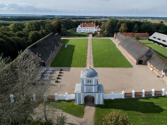 Luftfoto af en stor ejendom med et hvidt palæ, symmetriske græsplæner, to lange murstensbygninger og en udsmykket port i forgrunden, omgivet af træer og landskab.