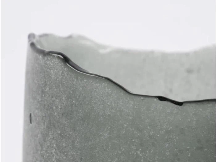 Et nærbillede af en gennemsigtig, struktureret glas- eller keramikkop med en ujævn, bølget kant mod en almindelig baggrund.