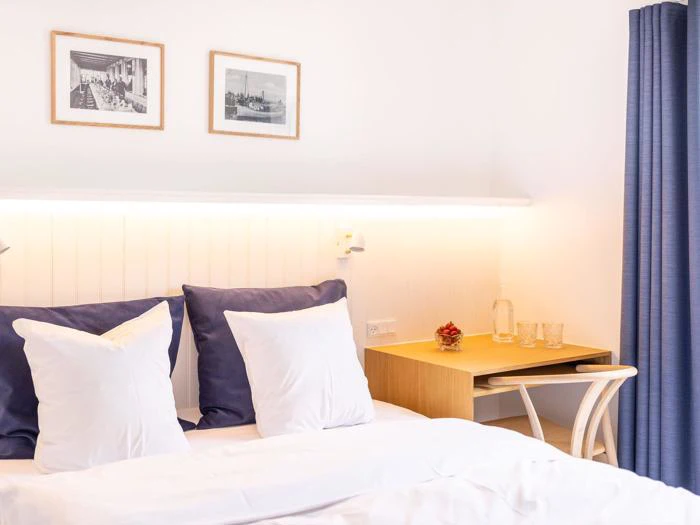 Et lyst, minimalistisk soveværelse med en seng, blå og hvide puder, et lille træbord med to glas og en flaske og to indrammede fotos på væggen ovenover.