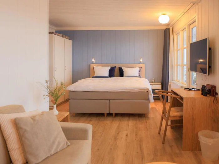 Hyggeligt soveværelse med en stor seng, blå accentvæg, klædeskab, skrivebord med stol, væghængt tv, lænestol og trægulv. Varm belysning og store vinduer skaber et lyst og indbydende rum.