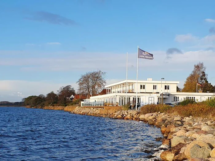 En hvid bygning med to flagstænger står ved siden af en klippekyst under en klar blå himmel med roligt vand i forgrunden og træer i baggrunden.