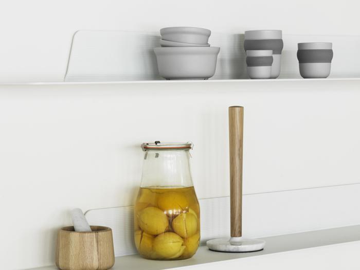 En køkkenhylde med stablede grå skåle, stribede kopper, en krukke med konserverede citroner, en morter og støder i træ og en køkkenrulleholder med træstang og marmorsokkel.