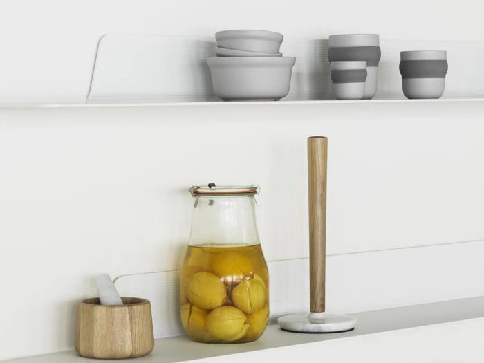 En minimalistisk køkkenhylde med en krukke med konserverede citroner, en køkkenrulleholder i marmor, en morter og støder samt stablede grå skåle og kopper på en hvid baggrund.