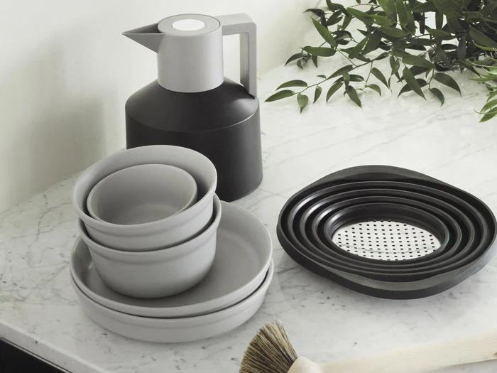 Minimalistisk køkkenudstyr på en marmorbordplade, inklusive stablede grå skåle og tallerkener, en sort og grå kande, indlejrede dørslag, en børste og grønne blade i hjørnet.