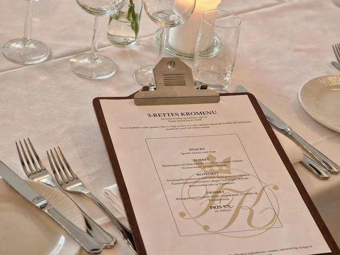 En udklipsmenu på en hvid dug, omgivet af bestik, glas, et tændt stearinlys og en brødtallerken ved en formel borddækning.