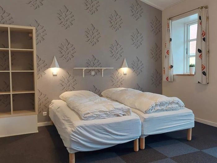 To enkeltsenge med hvidt sengetøj side om side i et moderne værelse med mønstret tapet, væglamper, en hylde, et vindue med gardiner og en åben reol.