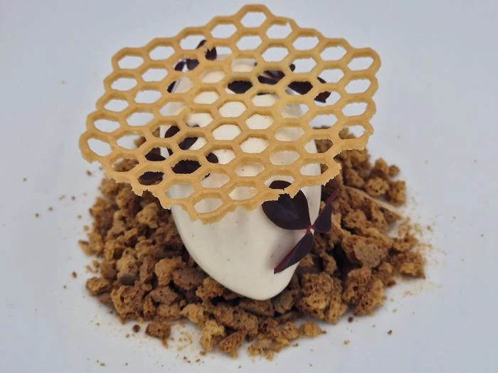 En kugle is toppet med en dekorativ honningkagemønstret vaffel, pyntet med små blade, ligger på en bund af smuldret brun kage.
