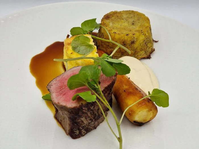 En gourmetret med en skive medium-rare oksekød, kartoffelkage, ristet pastinak, flødesauce, brun sauce og mikrogrønt som garniture på en hvid tallerken.