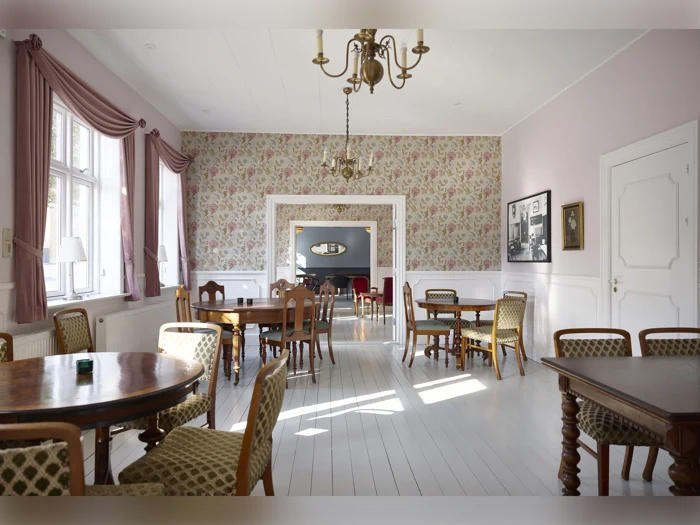 Et lyst, elegant rum med runde træborde, mønstrede stole, store vinduer, lysekroner, blomstret tapet og indrammet kunst på væggene. Sollyset strømmer ind fra venstre.