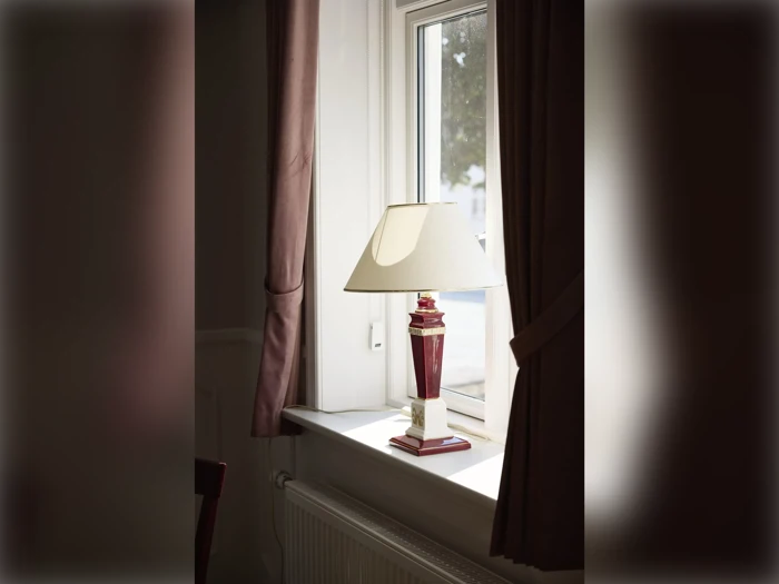 En rød og hvid bordlampe med en cremefarvet skærm står i vindueskarmen mellem to brune gardiner, og der kommer dagslys ind gennem vinduet.