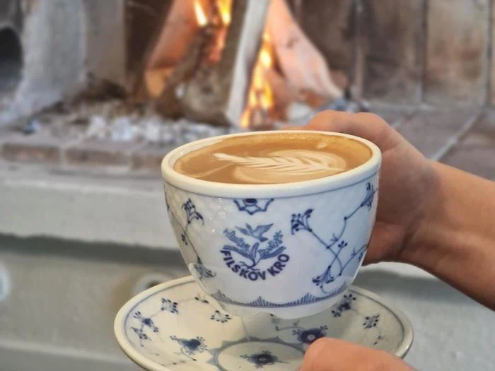 En person holder en blå og hvid mønstret kop latte med lattekunst foran en hyggelig pejs med ild i.