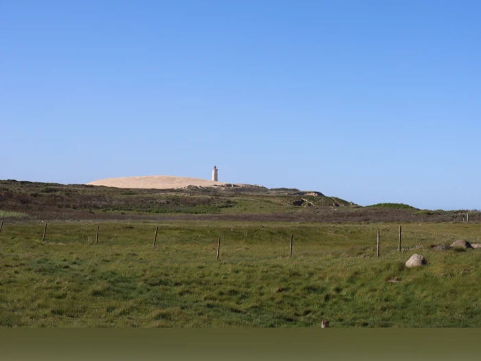 Et højt fyrtårn står på en sandbakke under en klar blå himmel, med en grøn græsmark og et hegn i forgrunden.