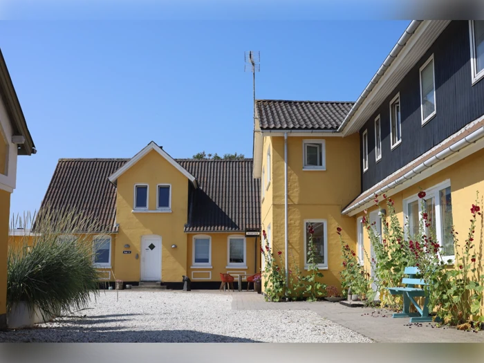 En solrig gårdhave med gule huse, hvide grusstier, grønne planter og en blå bænk. Himlen er klar og blå.
