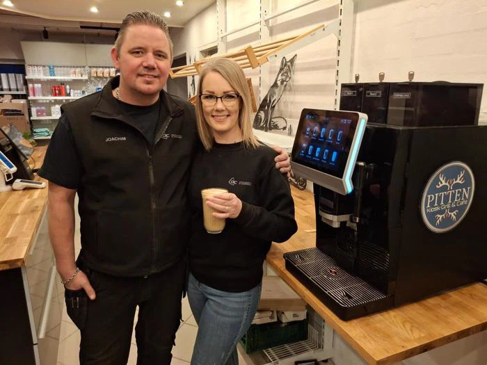 To personer står smilende ved en kaffemaskine på en trædisk; den ene holder en kop kaffe. Hylder og en ulveillustration er synlige i baggrunden.