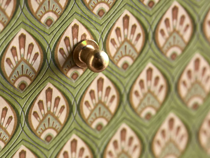 Nærbillede af en messingnål på et grønt og beige mønstret tapet med geometriske og blomstrede mønstre.