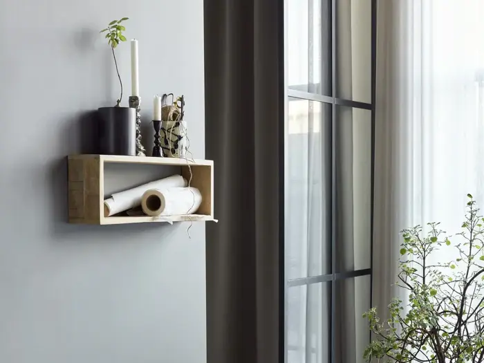 En lille væghylde af træ med stearinlys, en vase og sammenrullede papirer hænger på en grå væg ved siden af høje vinduer med gennemsigtige gardiner og en bladplante i nærheden.