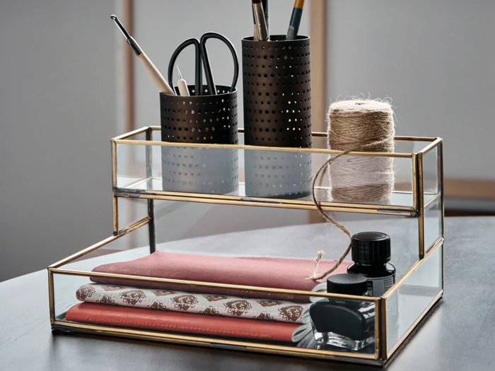 En organizer i glas og messing rummer notesbøger, mønstret papir, blækflasker, en garnrulle, sakse og kuglepenne i sorte holdere på en mørk bordplade.