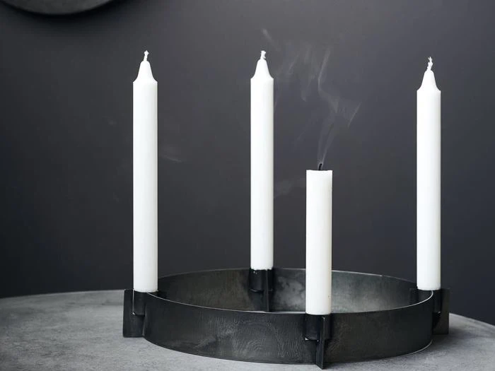 En rund, sort lysestage med fire hvide lys på en grå flade; det ene lys er slukket, og der stiger røg op fra vægen.