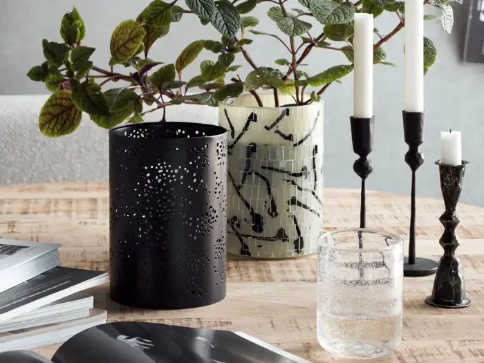 Et træbord med en sort udskåret lanterne, en mønstret vase med grønne bladstilke, tre lysestager med hvide stearinlys, et glas vand og åbne magasiner.