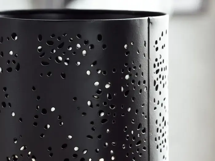 Nærbillede af en sort metalcylinder med mange uregelmæssigt formede huller, der skaber et dekorativt perforeret mønster.