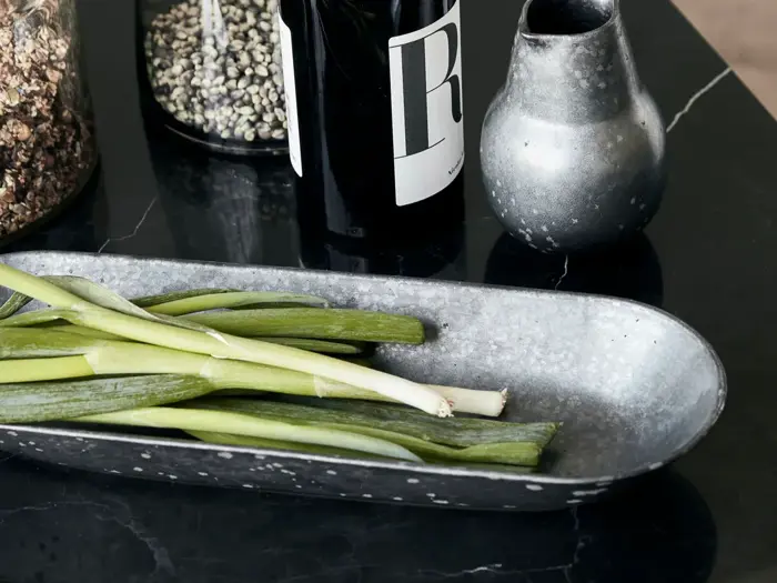 Grønne løg på en plettet grå bakke ved siden af en vinflaske, en lille metalvase og krukker med krydderier på en bordplade af sort marmor.