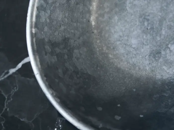 Nærbillede af en rund, forvitret metalskål med en spættet, grå overflade, placeret på en mørk, marmoreret baggrund.