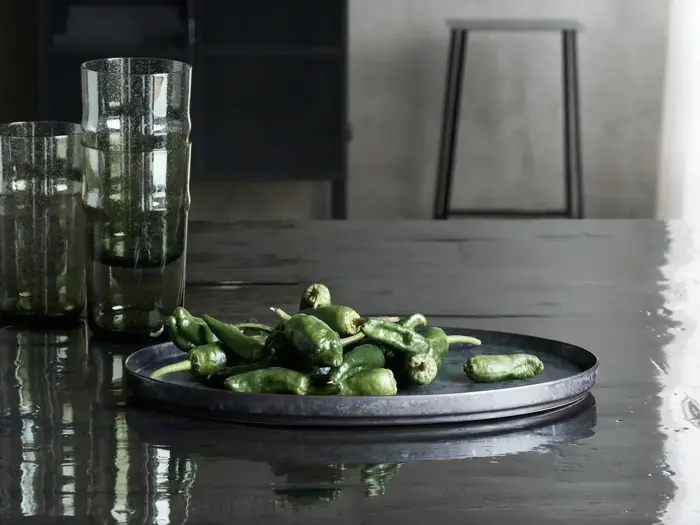 En metalbakke med friske grønne peberfrugter står på et blankt, mørkt bord ved siden af flere høje, røgfarvede glasbægre. En skammel og en sløret baggrund er synlige.