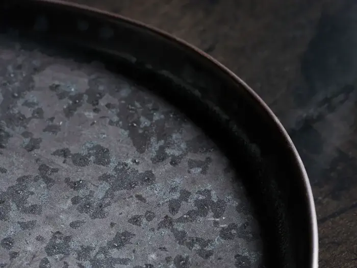 Nærbillede af en rund, mørk metalbakke med en plettet, forvitret tekstur, placeret på en mørk træoverflade.