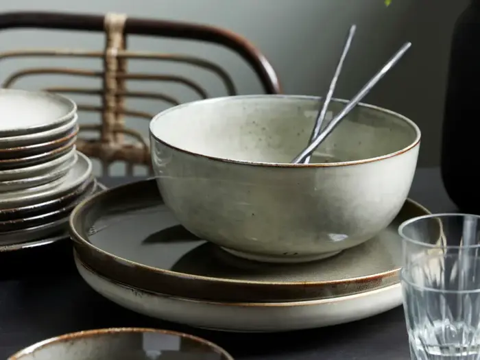 En keramikskål med to spisepinde i metal hviler på stablede tallerkener ved siden af en glaskop og flere tallerkener, med en stol i baggrunden.