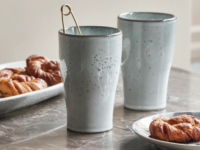 To keramikkopper med lysegrå glasur står på et marmorbord; i den ene kop ligger en guldske. Tallerkener med kager er i baggrunden og forgrunden.