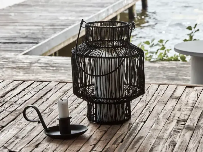 Et hvidt lys i en sort trådlygte og et andet lys i en sort metalholder står på et rustikt træbord ved en forvitret havn over vandet.