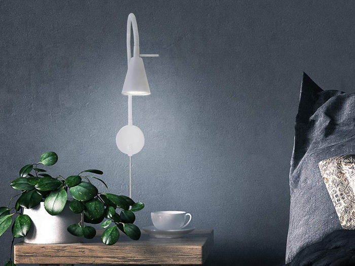 En moderne sengescene med en hvid væglampe, et træbord med en potteplante og en hvid tekop ved siden af en mørk pude.