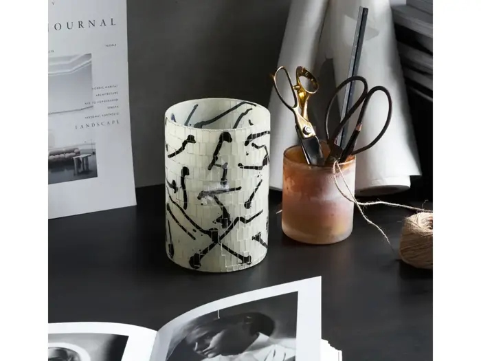 Et skrivebord med en hvid og sort vase med abstrakt mønster, en kop med sakse og blyanter, en spole med garn og åbne magasiner eller bøger.