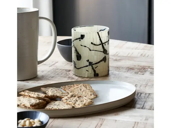 Et træbord med en tallerken med kiks, en skål med nødder, et keramikkrus, en lille mørk skål og en dekorativ lysestage med et sort abstrakt mønster.