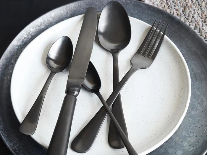 Et sæt matsort bestik - to skeer, en kniv og en gaffel - arrangeret på en hvid tallerken med en grå kanttallerken nedenunder.