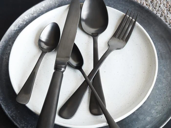 Et sæt matsort bestik med kniv, gaffel og to skeer arrangeret på en hvid tallerken med en mørkegrå oplader under.
