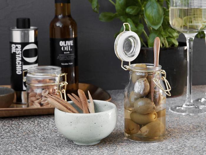 En krukke med grønne oliven med en træske, en lille skål med træskeer, nødder i en krukke, olivenolieflasker og et glas hvidvin på en plettet bordplade.