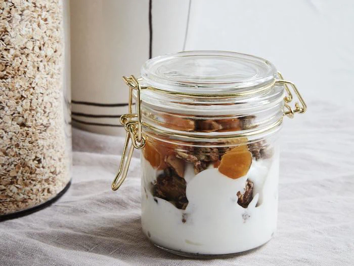 Et glas fyldt med yoghurt, granola og tørret frugt står på et bord ved siden af en beholder med havregryn.