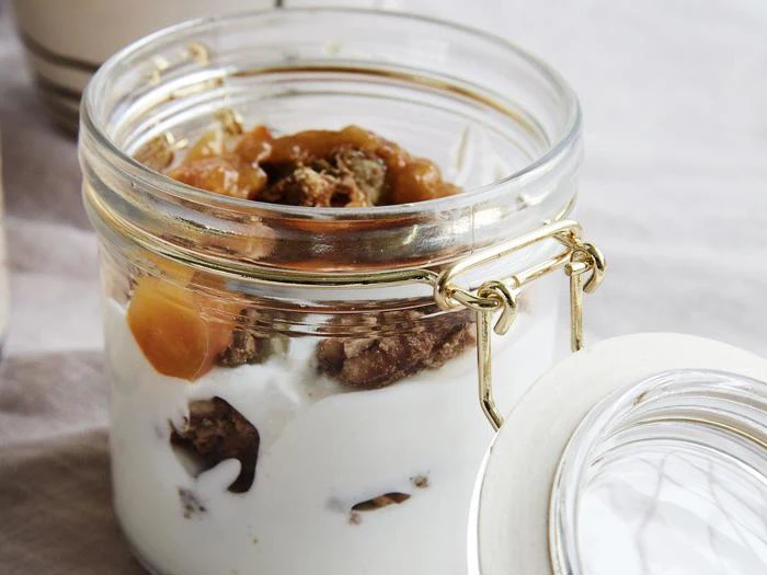 En glaskrukke fyldt med lag af cremet yoghurt, granola og frugtkompot, med låget åbent ved siden af på en lys overflade.