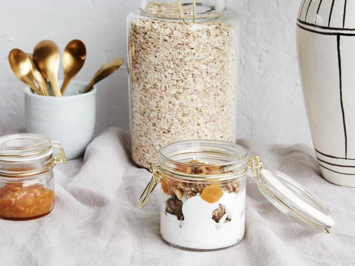 Et glas yoghurt med granola og frugt står på et bord. I nærheden står syltetøjsglas og havregryn, guldskeer i en kop og en hvid vase med sorte streger.