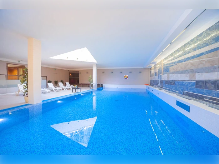 Indendørs swimmingpool med lyseblåt vand, hvide liggestole langs den ene side og en moderne flisebelagt væg. Ovenlysvinduet lukker naturligt lys ind.
