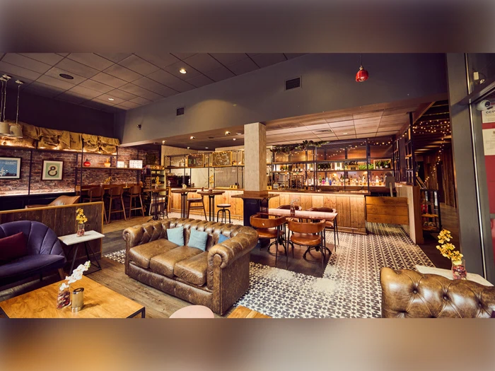 En hyggelig, stilfuld bar med lædersofaer, træborde, mønstrede flisegulve og en veloplyst bardisk i baggrunden. Rummet har en varm belysning og en moderne, rustik indretning.