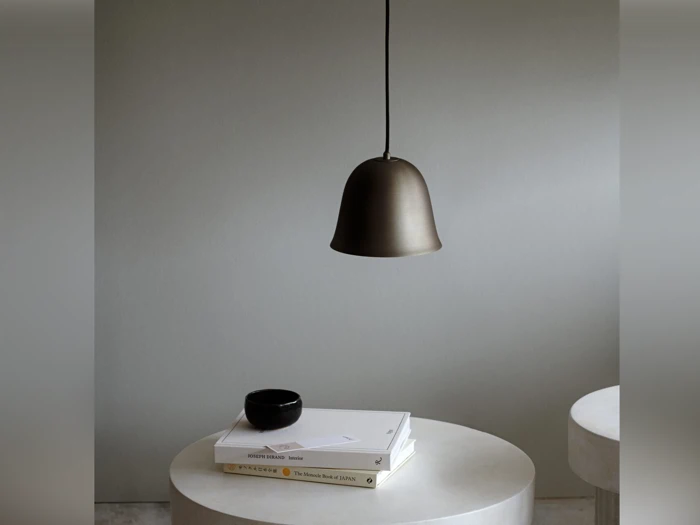 En minimalistisk scene med en bronzependel over et rundt hvidt bord med bøger, en lille sort skål og en seddel; et andet rundt bord er delvist synligt.