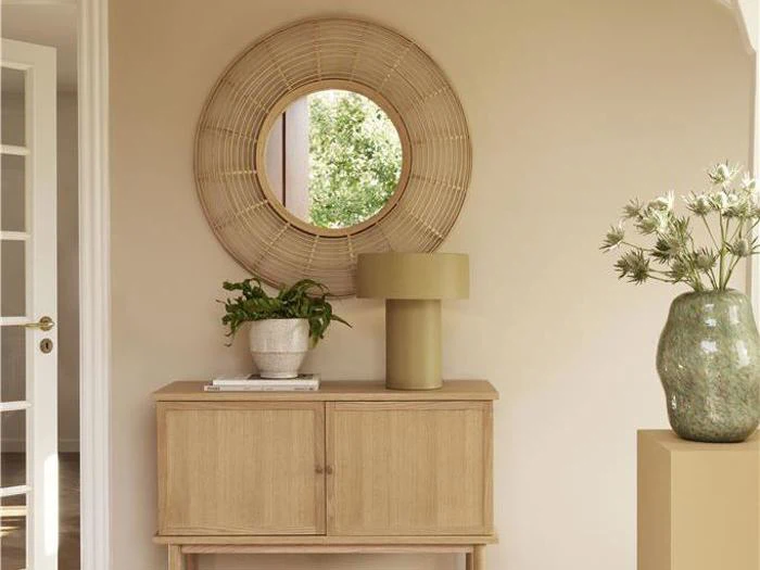 Et træskab med en lampe, en plante og en bog står under et rundt rottingspejl på en beige væg; en stor vase med blomster står til højre.