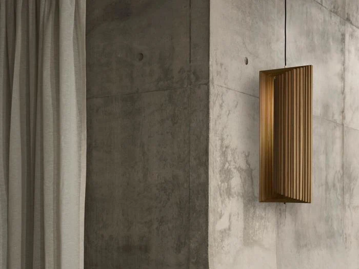 En guldfarvet væglampe med lodrette lameller monteret på en betonvæg ved siden af lysbeige gardiner i et minimalistisk interiør.