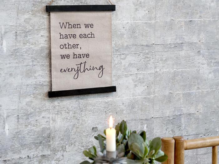 På et skilt på en grå væg står der: Når vi har hinanden, har vi alt. Nedenfor ses et tændt stearinlys og en grøn plante i forgrunden.