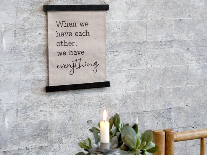 På et skilt på en grå væg står der: Når vi har hinanden, har vi alt. Nedenfor ses et tændt stearinlys og en grøn plante i forgrunden.