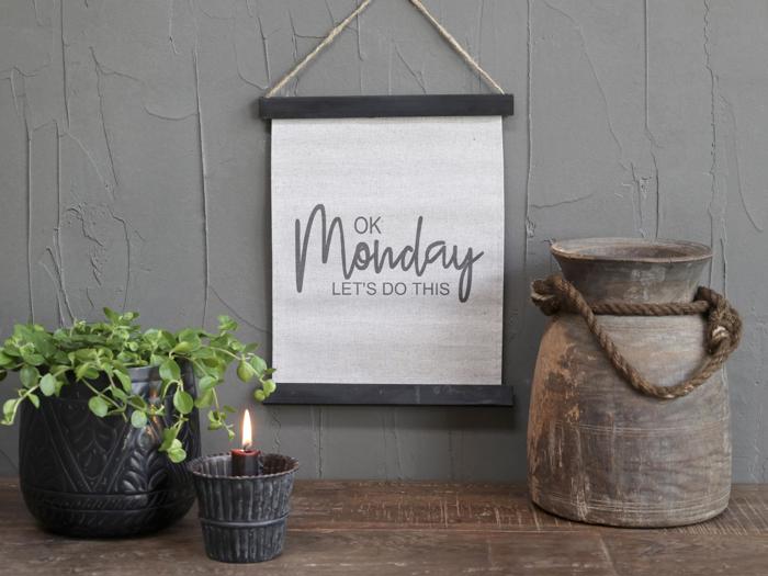 På et vægophæng står der OK Monday LETS DO THIS ved siden af en potteplante, et tændt stearinlys og en rustik trækande på en træoverflade.