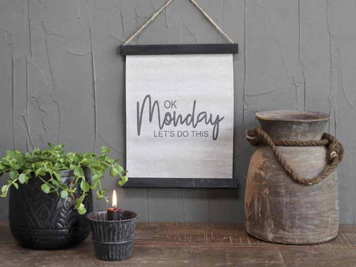 På et vægophæng står der OK Monday LETS DO THIS ved siden af en potteplante, et tændt stearinlys og en rustik trækande på en træoverflade.